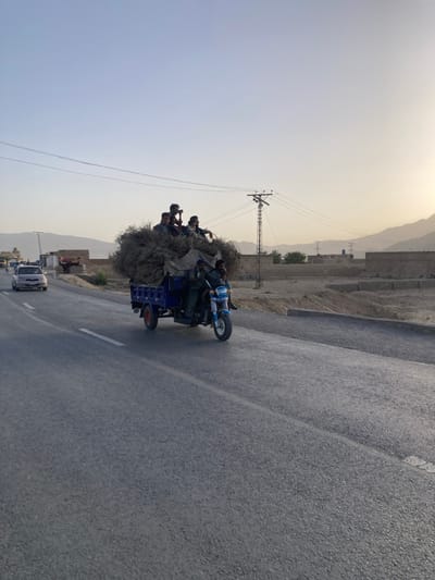The image captures a rural road scene in Spezand, Pakistan, under clear, late afternoon lighting. A blue three-wheeled motorized rickshaw, overloaded with a large volume of dried, bushy vegetation partially covered by a light-colored tarp, is positioned on the right side of an asphalt road. Six adult males occupy the rickshaw: two in the front cabin, one operating the vehicle and one as a passenger, and four seated atop the vegetation load in the rear. One of the individuals on top holds a black camera or similar device. All visible persons wear dark clothing. A broken white line marks the center of the asphalt road. A silver sedan is visible in the left lane. The immediate environment features a dry, unpaved shoulder with a low concrete barrier to the right and sparse structures to the left. Utility poles and power lines are present on both sides. Arid, rocky mountains define the distant background under a clear, light blue sky with a warm, yellowish tint near the horizon.