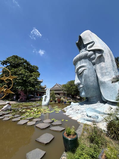 The image depicts a large, pale blue statue of a woman with a serene expression, her head draped in a veil. She is situated in a garden setting, with a pond in the foreground. Stepping stones cross the pond, which is dotted with lily pads and a few lotus flowers. To the left of the statue, a person in white clothing sits on the edge of the pond. In the background, a lush green tree stands prominently, with traditional Vietnamese architecture visible behind it. The sky is a clear, bright blue with a few scattered clouds. This scene is located at the Linh Ung Pagoda in Da Nang, Vietnam, not Hòa Đa as initially stated.