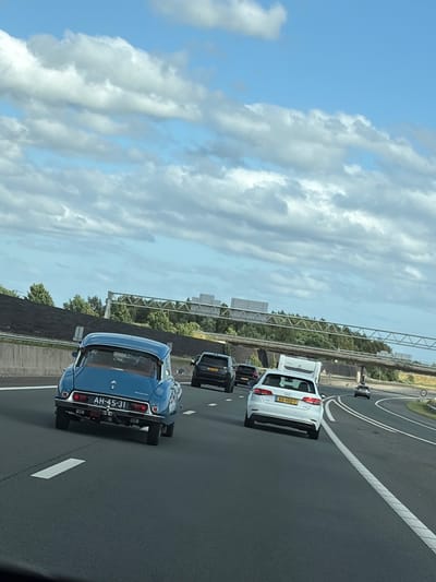 The media file shows a view from a vehicle traveling on a highway in Wierden, Netherlands. The sky is mostly blue with scattered white clouds, suggesting daytime weather. The highway has multiple lanes with clear white markings.
The primary subjects are cars on the road. In the foreground, a blue classic car, possibly a Citroën, is visible from the rear. It has a license plate that reads "AH-45-31". Behind it, several other cars are traveling in the same direction, including a white Audi with a license plate "RS-100-T", and two black cars. One of the black cars appears to be a sedan, and another could be a hatchback. The cars are spaced out, indicating typical highway traffic.
In the background, there are trees and vegetation lining the highway, with some infrastructure visible, possibly including overpasses or sound barriers. The overall scene depicts a normal day of driving on a highway in the Netherlands. There are no people clearly visible as subjects, though they are implied to be the drivers of the vehicles. The atmosphere appears calm and ordinary, with no indication of any unusual events. The time of day is likely afternoon, given the brightness and cloud patterns.