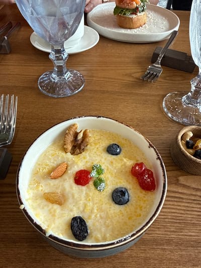 A high-angle, medium shot captures a wooden table set with breakfast items. In the foreground, a light-colored bowl filled with creamy, yellow porridge is topped with a walnut half, an almond, two blueberries, two red berries, mint leaves dusted with powdered sugar, and a few raisins. To the right of the bowl is a small wooden dish containing a mix of nuts and dried fruit, including cashews, raisins, and possibly cranberries. A fork rests to the left of the porridge bowl. In the background, a partially visible person's hands are visible reaching for a white plate holding a small open-faced sandwich with a red topping and greenery. Two clear, faceted glasses filled with water are also visible, one to the left and one to the right of the frame. The table surface is made of warm-toned wood planks. The setting appears to be a restaurant in Nizhnekayancha, Russia.