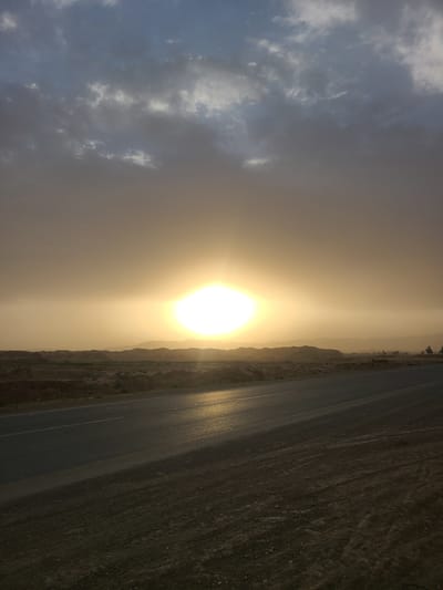 The image presents a wide, low-angle view of an arid landscape at either sunrise or sunset. A paved two-lane road, possibly an asphalt highway with a faint white dividing line, traverses the lower portion of the frame, appearing reflective from the intense light. Flanking the road is dry, rocky, and dusty terrain with minimal vegetation.
The most prominent feature is the extremely bright, circular sun, positioned in the middle-left of the horizon. It emits a powerful, golden glare that dominates the scene, creating a strong atmospheric haze and warm, golden-orange hues across the sky and landscape. This intense light suggests the sun is very low in the sky.
Above and to the far left, the sky transitions from soft, muted blue-grey clouds to the warm, hazy glow near the sun. In the distance, a range of dark, silhouetted mountains or hills forms a rugged horizon, partially obscured by the atmospheric conditions. Sparse, dark vegetation or small trees are barely discernible on the far right horizon, just above the mountains.
The scene evokes a sense of vastness and tranquility, characteristic of an open road traversing a desert or semi-desert region. There are no people or specific activities depicted, focusing solely on the natural environment. The notable details include the dramatic, atmospheric lighting of dawn or dusk, the reflective quality of the road surface, and the interplay of clouds, sun, and distant mountains in what appears to be a remote location. No visible text is present in the image.