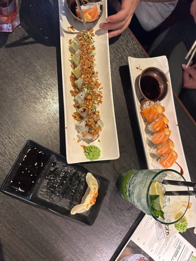 The image depicts a close-up, overhead view of a table at an Asian restaurant, likely in Leidschendam, Netherlands. The table is dark and wooden with a visible grain. On the table are two plates of sushi: one contains several pieces of sushi topped with crispy fried onions and green onions, and another plate features sushi rolls topped with salmon and a creamy sauce. Adjacent to the sushi is a small dish with soy sauce and a dollop of green wasabi. A black rectangular plate holds soy sauce in one compartment and a single, golden-brown gyoza in another, with some dark, shredded material. To the right, a tall, clear glass is filled with ice, mint leaves, and lime wedges, with two black chopsticks resting inside. A menu is partially visible at the bottom right. Two pairs of hands are visible, one holding chopsticks and reaching for sushi, and another partially obscuring a white shirt. The lighting suggests an indoor setting.
