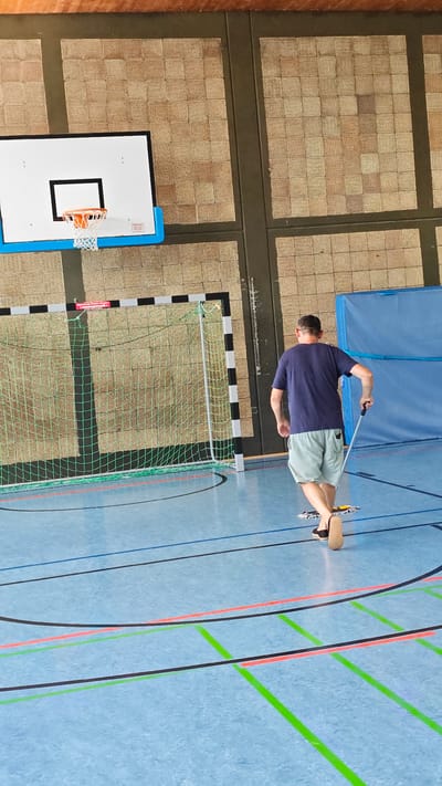 The image depicts a man mopping the floor of a gymnasium in Maintal, Germany. The man, seen from behind, is wearing a dark blue t-shirt and light-colored shorts. He is using a mop with a long handle and a flat cleaning head. The gymnasium floor is a light blue color with various colored lines indicating different sports courts, including red, green, and black lines.
In the background, a handball goal with a green net is visible. Above and to the left of the goal, a basketball hoop with a white backboard and an orange rim is mounted on the wall. The walls of the gymnasium are covered with a textured, beige material arranged in a grid pattern, typical of gymnasium interiors. There is also a large blue matting against the far wall.
The lighting suggests it is daytime, with natural light possibly coming from windows not visible in the frame. No specific weather conditions can be inferred. The activity is maintenance work, cleaning the sports facility. There is no obvious emotional expression conveyed due to the perspective and focus on the activity.
A small red and white sign is visible on the back of the handball goal, but the text is illegible. The overall scene is one of a typical indoor sports environment undergoing cleaning.