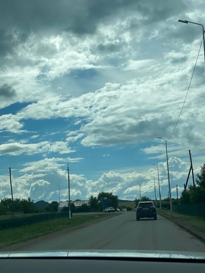 A grey SUV is driving down a paved road in a rural area, possibly in the city of Şalqar, Kazakhstan. The road is flanked by grassy verges and a dark green fence. Trees and buildings are visible in the distance. The sky is a dramatic mix of dark grey and bright blue clouds, with several tall light poles lining the road.