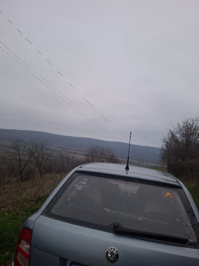 A light blue Skoda hatchback, identifiable by its rear emblem, is positioned on a grassy embankment in Balkantsi, Bulgaria. The vehicle's rear window features a yellow triangular warning sticker with a black exclamation mark on the right side and an indistinct light-colored sticker on the left. A black roof-mounted antenna is present above the rear window. The environment beyond the vehicle comprises a rural landscape with rolling hills, sparse bare deciduous trees, and visible patches of cultivated land. The sky is uniformly grey and overcast. Two sets of overhead utility lines, one appearing solid and the other dashed, traverse the upper frame diagonally from left to right. No individuals, specific actions, or interactions are discernible.