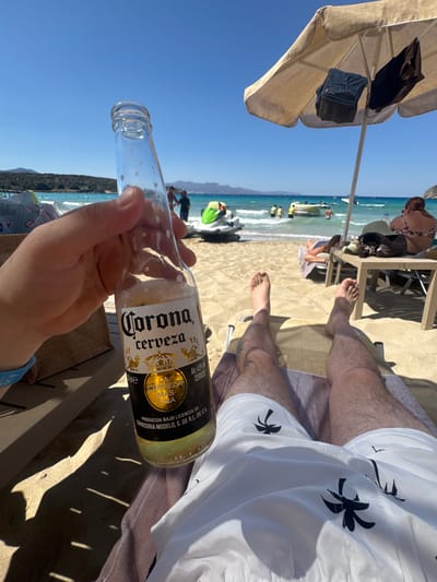 The image captures a sunny day at a beach. The foreground is dominated by a person's hand holding an almost empty bottle of Corona beer, with some condensation visible on the glass. The bottle's label clearly reads "Corona Extra Cerveza" and provides details about its alcohol content and production. In the background, the turquoise water of the sea stretches out, with a few jet skis visible in the distance. People are scattered on the sandy beach and near the water's edge, some appearing to be relaxing or engaging in water activities. A beige beach umbrella with clothing items draped over it provides shade. The overall atmosphere is one of leisure and vacation, typical of a beach setting on a bright, clear day. The clear blue sky suggests good weather conditions. The location appears to be a coastal area with a view of mountains in the distance.