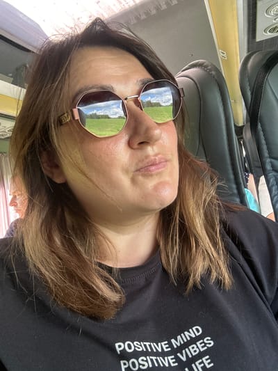 In a medium close-up shot, a woman with shoulder-length brown hair and a light complexion is positioned in the foreground, looking slightly to her right. She is wearing large, hexagonal sunglasses with rose-gold frames, which reflect a green field under a cloudy sky. Her mouth is slightly open, and her expression is neutral. She is wearing a black t-shirt with white text that reads "POSITIVE MIND POSITIVE VIBES LIFE." Behind her, the interior of a bus is visible. The seats are upholstered in dark gray leather. In the background, to the left, another person with reddish-blonde hair is partially visible, looking out a window. The location is specified as Ivanovska, Russia. The scene appears to be captured during daylight.
