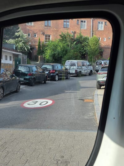 The image, taken from a car's side mirror, shows a street scene in Sopot, Poland. In the foreground, a red and white circular speed limit sign indicating "30" is painted on the asphalt. Several cars are parked along the street: a dark green hatchback, a black sedan, a white van with faded paint and graphics, a silver minivan, and a light gray hatchback. The background features a multi-story brick building with numerous windows and trees bordering the property. The street is paved, with a sidewalk made of interlocking stones. The lighting suggests it is daytime.