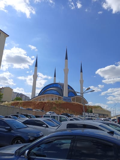 The image shows the Bayraktar Mosque in Yahşihan, Turkey. The mosque features a large blue-domed roof and four minarets. In the foreground, a parking lot is filled with various cars. The sky is a bright blue with scattered white clouds. The mosque is situated on a hillside with a backdrop of residential buildings. A street lamp is visible in the middle of the frame. There are no people clearly visible in the image.