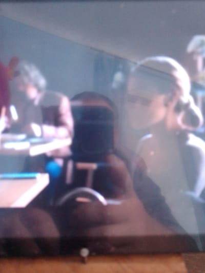 The image shows a dimly lit, slightly blurry interior scene. Several people are visible, mostly out of focus. In the foreground, on the left, a person is partially visible with a pinkish hue on their left arm. To their right, a dark, upright object that resembles a monitor or screen is present. In the mid-ground, a person with light-colored hair styled in a bun is looking to the right, and their face is illuminated. To the left of this person, another individual's head and shoulder are visible. The background is indistinct, with some lighter areas that could be walls or other surfaces. There are horizontal surfaces in the scene that appear to be tables or desks, with some lighter-colored objects on them. The overall impression is of an indoor gathering or workspace. The image does not contain any discernible information that would indicate a specific location in the city of Kotiv, Ukraine.