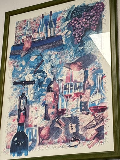 This is a framed watercolor painting depicting a still life composition related to wine. The artwork features several elements including bottles of wine, wine glasses filled with red wine, grapes (both purple and white varieties), and wine corkscrews. There are also what appear to be snails incorporated into the design. The background has a textured, abstract quality with some visible script or writing. The painting is signed by "KLAUS D. SCHIEFFLER" and dated "1996". The framing is a simple green wood. The image of the painting is taken from an angle, looking slightly down and to the right, with a white piece of paper or matting partially obscuring the top right corner of the artwork and creating a glare on the glass. There is no information in the image or its crops that indicates the location of this painting within the city of Mülheim an der Ruhr, Germany.