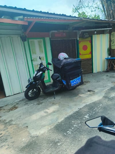 The image shows a black motorcycle parked on a street in Sukasari, Indonesia,  at the coordinates -6.837, 107.598.  The motorcycle is a delivery vehicle with a black bag attached to the back, bearing the words "WOW SPACE IT". The motorcycle is parked in front of a building with a green and white striped wall and a wooden door. There is a sign on the building with the words "Siap Siap" in Indonesian. The building is next to a second building with a yellow door. The door is decorated with a large red circle. Both buildings are in a state of disrepair. The street is paved and covered with a layer of dirt. There is a small plant in the corner of the image, next to the yellow door.  The motorcycle is empty, and there is no one visible in the image.  The camera is positioned at a low angle, looking up at the motorcycle and buildings.  The photograph seems to be taken from another vehicle.