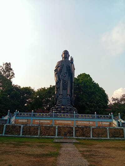 A large, dark Buddha statue stands prominently in Phú An, Vietnam. The statue is set upon an elevated platform that features carved panels with illustrations. The platform's upper section has signs with text written in Vietnamese and Chinese characters. A stone path leads directly towards the statue from the viewer's perspective. Trees form a backdrop behind the statue and its platform. The sky is a gradient of light blue near the top, transitioning to a hazy, warmer tone closer to the horizon.