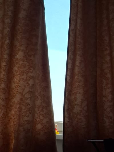 The image depicts a view through partially drawn, patterned brown curtains, revealing a bright blue sky and a sliver of a window frame. On the windowsill, a few colorful toy blocks and a small figurine are visible. The scene appears to be an interior view looking outwards, suggesting a residential setting within the city of Ilava, Slovakia. The lighting indicates it is daytime.