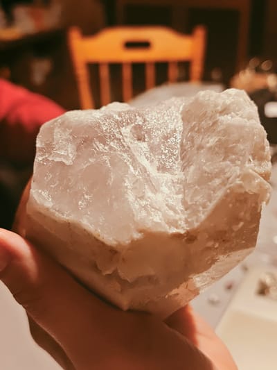 A hand holds a large, roughly cubic piece of translucent white mineral, possibly quartz. The bottom portion has a more opaque, beige hue. The background is out of focus, but includes a wooden chair with a cutout back, a red article of clothing, and other indistinct objects. The location is unspecified and therefore cannot be confirmed as Ильинское, Russia.