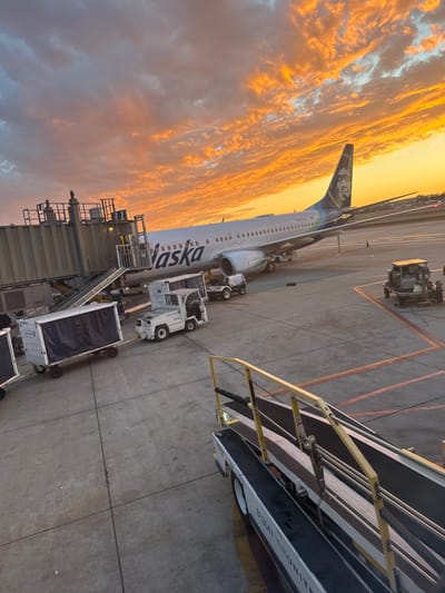 The image displays an airport tarmac at John Wayne Airport (SNA) in Santa Ana, California, United States, during sunset. A white Alaska Airlines Boeing 737 aircraft, tail number N222AS, is parked at a gate, with a gray jet bridge connected to its forward port side. The sky above is filled with prominent orange and yellow stratocumulus clouds. On the concrete apron, multiple pieces of ground support equipment are present: a train of covered baggage carts, marked "UNITED COACH," is positioned near the aircraft's forward cargo hold. A white aircraft tug, also labeled "UNITED COACH," is connected to these carts. In the foreground, a portion of a belt loader with yellow railings and "BL3547 TUE UNITED" visible on its side is observed. Another United-branded service vehicle is located near the aircraft's aft section. No individuals are visible. Runway or taxiway lighting is discernible in the distant right background.