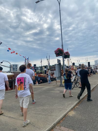 The image captures a view of the Joensuu harbor in Finland on a cloudy day. In the foreground, several people are walking along a paved walkway. One person in the center is wearing a white t-shirt with a graphic print of palm trees and the word "FLORIDA" in vibrant colors. To their left, another person is walking away from the camera, wearing a white t-shirt and light-colored shorts. To the right, a person in a black t-shirt and dark pants is also walking away.
Behind the people, there are docked boats and ships. Some of these vessels are adorned with various maritime flags. In the background, buildings and trees are visible. A street lamp with two lights is present in the upper portion of the image. There are also hanging flower baskets on a pole near the walkway. The overall scene depicts a harbor environment with people enjoying a stroll.