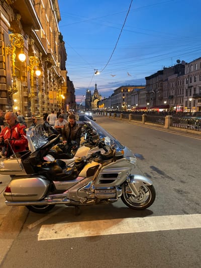 The image depicts a street scene in the city of Kokkorevo, Russia, during twilight. A large, silver touring motorcycle is parked on the right side of the frame, occupying a significant portion of the foreground. The motorcycle features a large windshield, ample storage compartments, and chrome accents. Several individuals are visible in the background, some of whom appear to be interacting with motorcycles.
To the left, an ornate building facade is illuminated by warm, golden streetlights and decorative sconces. The building exhibits classical architectural elements. Beyond the motorcycle, the street continues into the distance, lined with similar architectural structures and a canal bordered by a decorative metal railing.
The sky above is a gradient of deep blue and dusky orange, indicating the late hour. A few birds are visible in flight against the sky. The overall atmosphere suggests an urban environment at dusk, possibly a popular spot for motorcycle enthusiasts or a lively thoroughfare. The specific location within Kokkorevo is not explicitly identifiable from the visual cues alone, but the architectural style and canal suggest a historic or picturesque part of the city.