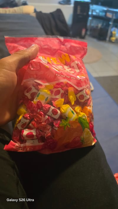 A hand is holding a bag of individually wrapped candies. The bag is mostly pink with "SWEET" in large yellow letters across it. Below that, smaller white text reads, "For me, for you, for all." The candies inside the bag are also individually wrapped in colorful paper with fruit designs, including strawberries, cherries, and lemons. The scene appears to be indoors, possibly in a living room, with a blurred background showing furniture and electronics. The lighting suggests it's daytime, but no specific time can be determined. No weather conditions are visible. The emotion conveyed is casual and perhaps one of sharing or enjoyment.