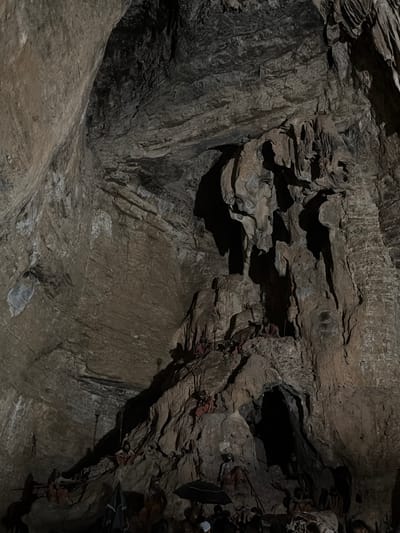 The image depicts the interior of a cave, likely Yaganti Caves, in Bethamcherla, India. The cave walls are rough and textured, with stalactites and stalagmites visible. Several figures, appearing to be effigies or statues dressed in red and white fabric, are arranged on ledges and within the cave's natural formations. Some figures are seated, while others are standing. The lighting is dim, with some areas casting deep shadows, suggesting an artificial light source illuminating the scene. The overall impression is one of a sacred or historical site within a natural cavern.