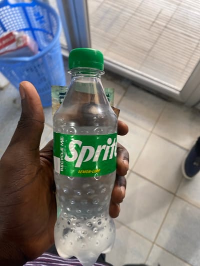 The image displays a close-up view of a dark-skinned hand holding a clear plastic Sprite bottle horizontally. The bottle has a vibrant green screw cap and a green label with the prominent "Sprite" logo in white. Below the main logo, the words "LEMON-LIME" are visible, and "RECYCLE ME!" is printed towards the bottom of the label. The bottle's surface features a textured pattern of small, circular indentations.
Inside the empty plastic bottle, several banknotes are visible, folded and tucked into the bottle's opening. One note clearly shows the number "10," indicating a denomination. Given the context of Goderich, Sierra Leone, these are likely Sierra Leonean Leone (SLL).
The background suggests an indoor setting, possibly a shop or market. A light-colored tiled floor with visible grout lines and some reflections is prominent. On the left side of the frame, a blue, mesh-patterned basket or shopping cart is partially visible. Further in the background, a blurred dark shoe or foot can be seen on the floor. The scene is illuminated by what appears to be ambient indoor lighting. No specific activity beyond holding the bottle is depicted, and no emotions are discernible.
