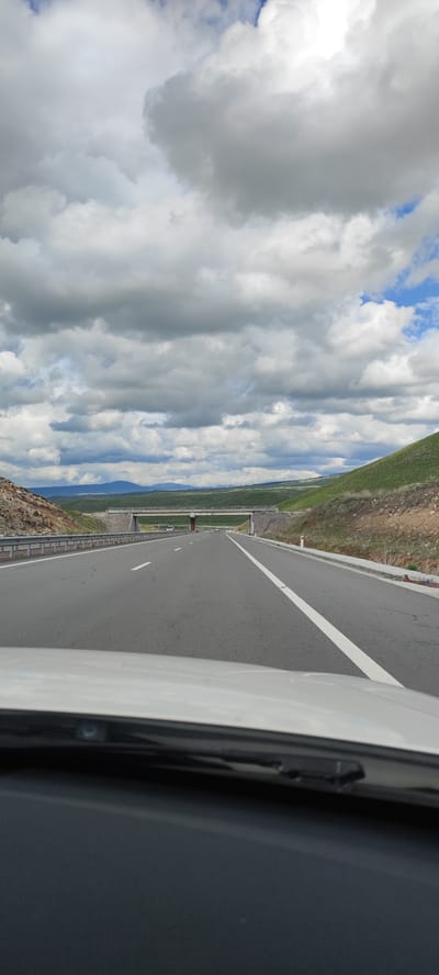 The image depicts a view from inside a vehicle traveling on a highway in a rural area. The road is paved with two lanes going forward, indicated by solid white lines on the right side of the road and a dashed white line on the left. An overpass structure is visible in the distance, spanning across the highway. The landscape on either side of the road consists of hilly terrain, sparsely covered with green vegetation. The sky is cloudy with patches of blue visible. There's no explicit confirmation of the location being Dzorakap, Armenia from the image. No people are visible, and the focus is on the road, landscape, and sky.