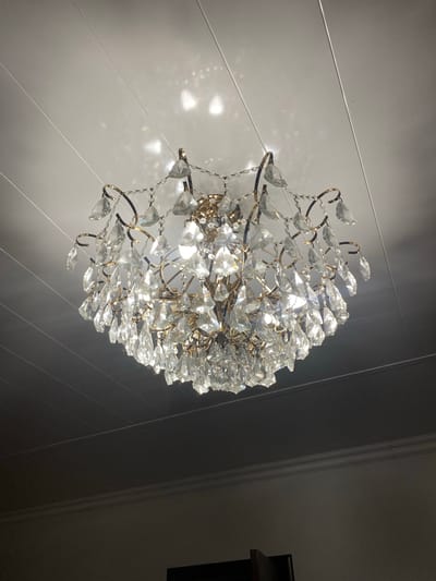 A crystal chandelier hangs from a white, paneled ceiling. The chandelier is ornate, with multiple tiers of clear crystal pendants and a gold-toned metal frame. Light reflects off the crystals, casting dappled patterns on the ceiling. The ceiling is divided by thin, horizontal lines, suggesting a suspended ceiling or paneling. Below the ceiling, the upper portion of a wall is visible, painted a dark, neutral color, with white crown molding separating it from the ceiling. The lower part of the image is dark, obscuring the rest of the room. There are no people or animals visible in the image, nor are there any obvious signs of activity or interaction. The scene appears to be an interior, likely a residential or formal space, given the style of the chandelier. The location within Gergebil, Russia, is not visually indicated by any specific landmarks or context within the image itself.