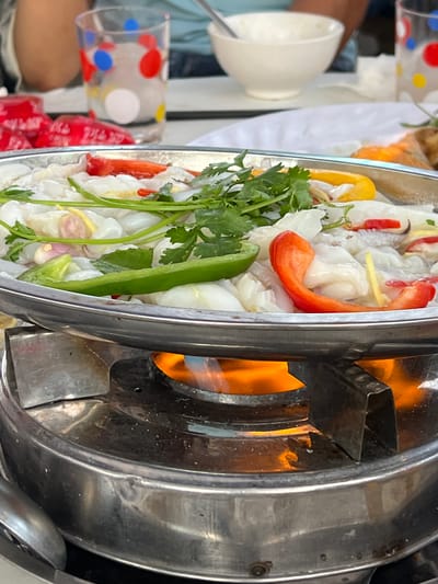 This vertical close-up image, captured in Bình An, Vietnam, showcases a metal cooking vessel filled with ingredients, heated by an active open flame from a burner below. The dish primarily consists of numerous translucent white pieces, possibly squid or jellyfish, artfully arranged and mixed with vibrant vegetables. Red and yellow bell pepper slices add contrasting color, alongside a long green chili pepper, fresh cilantro sprigs, and thin slivers of what appears to be ginger or shallots.

The flame under the pot emits a bright orange glow, indicating that the food is actively being cooked or kept warm. In the blurred background, a white ceramic bowl and a clear glass decorated with red, blue, yellow, and white polka dots are visible, suggesting a dining setting. A person's arm, clad in a light blue sleeve, is also partially seen. Further back, stacks of red-packaged items, possibly drinks or snacks, hint at a bustling food stall or market environment. The bright natural light suggests the scene takes place during the day. No explicit text is visible in the image.