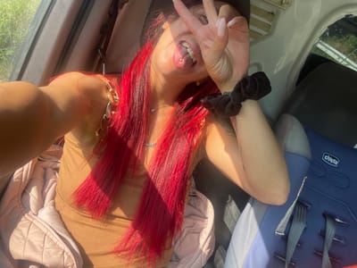In Zheglartsi, Bulgaria, a young woman with vibrant red hair, wearing a tan ribbed tank top and a pink puffer jacket, is captured in a selfie taken from inside a vehicle. Her tongue, adorned with a silver piercing, is visible as she flashes a peace sign with her right hand, obscuring part of her face. A black scrunchie is present on her wrist. The background reveals the interior of the car, including a blue child car seat with the "Chicco" logo and gray seatbelts. Sunlight streams into the vehicle, casting shadows and highlighting the scene.