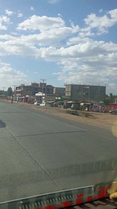 A street scene shows a flat, gray asphalt road receding into the distance under a bright sky dotted with white, puffy clouds. On the right side of the road, there are buildings, some appearing to be under construction, alongside businesses with signage including “HARDWARE”. Makeshift shelters and stalls are set up next to the buildings. People are walking along the side of the road. The scene seems to be captured from inside a vehicle with part of the vehicle's interior visible at the bottom.