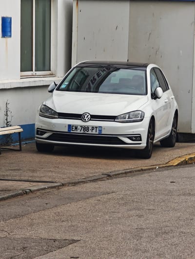 A white Volkswagen Golf, parked on a paved surface, is the primary subject of this image. The car has a black roof and its headlights and grille are clearly visible. The license plate of the car reads "EM-788-PT" and displays the European Union and French flag symbols, along with the number 62. Behind the car, there is a white building with a large window and a blue sign on the wall to the left. A simple wooden bench is situated to the left of the car. The ground is a rough, grey pavement with a yellow curb visible on the right side of the image. The lighting suggests it is daytime, with no strong shadows indicating a specific time. The scene appears to be an outdoor public space, possibly a parking area or a street, in Boulogne-sur-Mer, France. The overall impression is of a calm, everyday scene, with no discernible activity beyond the car being parked. The blue sign on the wall has some text, but it is not entirely legible.