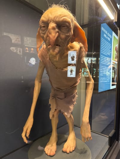 The image depicts a Dobby house-elf animatronic figure on display inside a glass case. A label is visible behind the figure with text that includes the words "HOUSE-ELVES" and other Japanese text. The setting appears to be an exhibit at the Harry Potter Studio Tour in Nerima, Japan. The figure is brown with large ears and eyes, and is dressed in tattered brown fabric. It is positioned on a gray platform inside the case. Light reflects off the glass case.