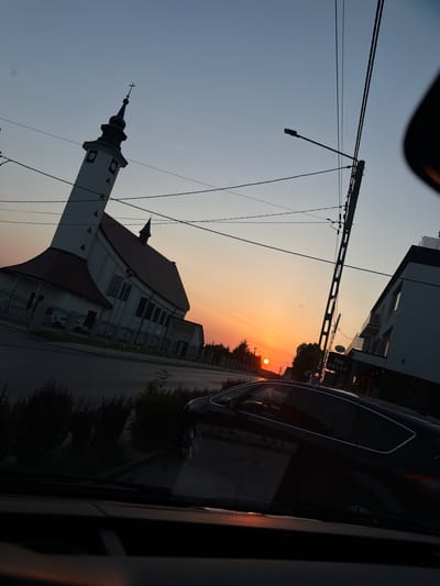 The image depicts a street scene in Mordarka, Poland, at sunset. The sun is low on the horizon, casting a warm, orange glow across the sky, transitioning to a pale blue at the top of the frame. A white church with a tall, dark steeple and a red roof is prominently featured on the left side of the image, partially silhouetted against the sky. The church has several square windows with clock faces visible on its tower.

In the background, modern buildings with dark facades and balconies are visible. A dark-colored car is parked on the right side of the street, with its reflection visible in the car's windows. The street is paved and lined with trees and bushes, which are mostly in silhouette. Electrical wires crisscross the sky, supported by utility poles.

The scene appears to be captured from within a vehicle, as indicated by the reflections and the partial view of the car's interior. The overall mood is serene and peaceful, typical of a sunset in a European town. No people are clearly visible in the image. One of the buildings in the background has text that appears to read "SKLEP," which is Polish for "shop."