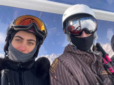 Two individuals in ski gear are depicted inside what appears to be a ski lift cabin in Valtournenche, Italy. The person on the left, a female, wears a black ski helmet, orange-tinted ski goggles pushed onto the helmet, a grey neck gaiter covering her lower face, and a black quilted ski jacket with fur trim. Her eyes are visible, directed towards the viewer. The person on the right, a male, wears a white ski helmet with a Rossignol logo, mirrored ski goggles reflecting a snowy slope and a person in red, and a black face mask covering his lower face. He is dressed in a brown ski jacket with thin white vertical stripes and a patch on the left sleeve. A black and pink ski pole is visible next to his right side. Both individuals are positioned from the chest up. The environment visible through the transparent cabin walls includes a clear blue sky and distant snow-covered mountains, indicating a sunny day.