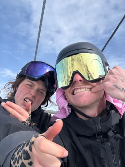 A close-up selfie depicts two individuals on a ski lift within the ski area of Le Monêtier-les-Bains, France. The subject on the left, identified as female, has reddish-brown hair and freckled skin. She wears a black ski helmet and dark goggles with a purple reflective lens. Her mouth is open with teeth visible in a grimace. Her left hand, partially covered by a leopard-print patterned garment, extends two fingers (index and middle) towards the camera. The subject on the right, identified as male, wears a black ski helmet, large gold-mirrored Oakley goggles, and a pink bandana visible around his neck/chin area. He has a light brown mustache. His left hand, which displays finger tattoos, manipulates the corner of his mouth downwards, exposing his upper teeth in a strained expression. Both individuals wear black ski jackets. A thick steel ski lift cable is visible diagonally across the upper portion of the frame, set against a partially cloudy blue sky.