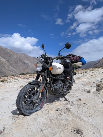 A Royal Enfield motorcycle is parked on a rocky, sandy surface with mountains in the background. The sky is blue with white clouds. The motorcycle is white with "Royal Enfield" written on the fuel tank. It has a black frame, black wheels, and a black seat. There are bags strapped to the back of the motorcycle. The license plate reads "LA-02A-5897". The scene appears to be in a remote, mountainous area, possibly in the vicinity of Sasoma, India, given the terrain and vegetation.