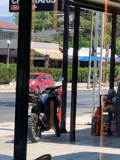 A street scene is captured from behind vertical dark poles, possibly a window frame, in Νέα Ραιδεστός, Greece. In the foreground, a portion of a black motorcycle with a prominent tire and exhaust is visible. To the left of the motorcycle, a red car is parked on the street. Behind the vehicles, a sidewalk is lined with green bushes, and beyond that, buildings with tiled roofs and some trees are present. Further in the background, there are structures with white awnings and blue and white coverings, suggesting commercial establishments or market stalls. Sunlight casts shadows on the tiled ground in the foreground. To the right, a display rack filled with sports balls, including soccer balls, is situated near an orange bag. A street lamp is visible on the left side of the image. The overall atmosphere appears to be a daytime scene on a street.