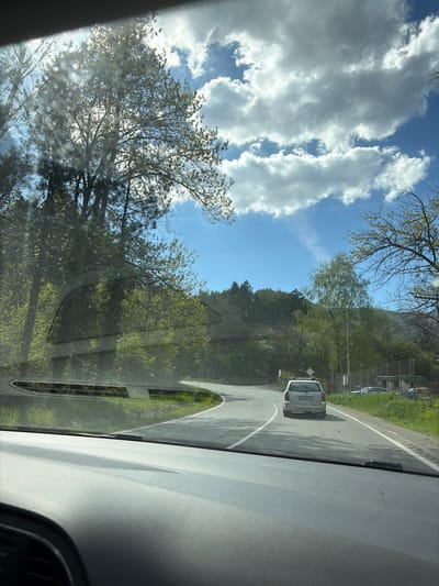 The image shows a view from inside a car, likely a driver's perspective. The car is driving on a paved road with a white line marking the edge. A silver car is visible ahead, and there is greenery, including trees and possibly a hillside, on either side of the road. The sky is blue with scattered clouds. The location may be Lukovo, Bulgaria based on the license plate of the silver car: CA 7389 KN. There are no visible people inside of the car or outside of the car that are discernible.