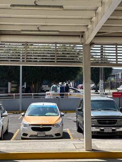 A parking area has several vehicles. A white and yellow taxi with plate number "CO-2835" and "REGION CORDOBA" displayed. A dark SUV with plate "YLB-096-B" is parked next to the taxi. People stand near a taxi dispatch area. The scene is partially framed by a white, covered walkway with a slatted design. A street sign and another vehicle are visible in the background, adding depth to the image.