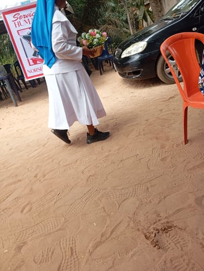 The image depicts an outdoor scene in Okpatu-Ukehe, Nigeria, on reddish-brown sand. A single individual, likely female, is captured mid-stride, moving from left to right. They are dressed in a white uniform consisting of a long-sleeved top and a pleated, calf-length skirt, paired with a bright blue head covering and black athletic shoes. The individual holds a light-brown woven basket containing a bouquet of artificial flowers with pink, white, and green elements. In the left background, a white banner is partially visible, displaying red text including "Service," "HUMAN," and "NORSE," alongside a graphic of a figure holding a staff. Several dark plastic chairs are positioned near the banner. In the mid-ground, to the right of the individual, a black sedan, possibly a Toyota, is parked, showing damage to its front bumper. An orange plastic chair is partially visible in the right foreground. The sandy ground exhibits numerous shoe prints. Green foliage and trees are present in the distant background, indicating an outdoor, vegetated setting.