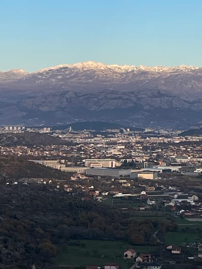 A panoramic, high-angle view depicts an urbanized valley landscape under a clear blue sky, extending towards snow-capped mountains in the distant background. The scene is located in Montenegro, overlooking the area encompassing Gornji Kokoti and the city of Podgorica.

The upper fifth of the image is occupied by a clear, light blue sky. Below this, a range of rugged mountains, dark in tone, exhibits patches of white snow on its peaks and upper slopes.

The mid-ground features a sprawling urban and suburban environment. In the further mid-distance, a denser city, identifiable as Podgorica, is visible with numerous high-rise buildings and a prominent white cable-stayed bridge (Millennium Bridge) spanning a river. Closer to the foreground, the terrain consists of rolling hills and valley floor, developed with a mix of residential structures, characterized by red and light-colored roofs, interspersed with green plots of land and scattered trees. This area represents the locale of Gornji Kokoti, extending into the suburban sprawl leading to Podgorica. Larger, rectangular commercial or industrial buildings with flat roofs are also visible within this mid-ground urban fabric.

The immediate foreground, occupying the bottom fifth of the image, shows dark, heavily vegetated hillside terrain, indicating the elevated vantage point from which the photograph was taken. A few isolated houses with visible roofs are situated on these lower slopes.

No individuals, specific actions, or interactions are discernible due to the distant, high-altitude perspective. The lighting suggests either late afternoon or early morning, casting long shadows across parts of the landscape and highlighting the snow on the distant mountains.