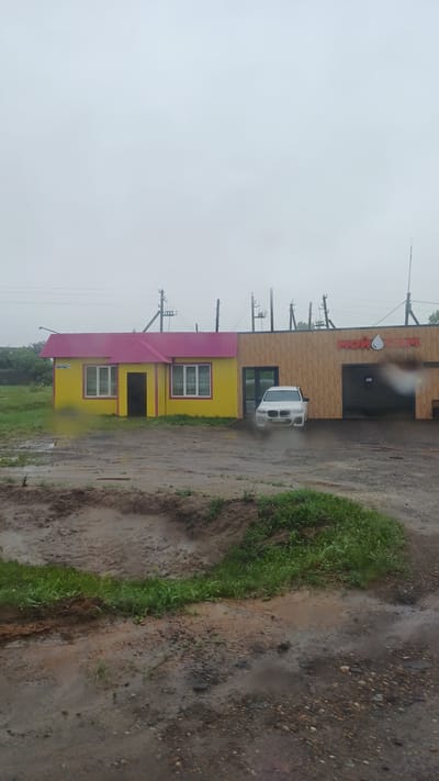 The image depicts a car wash in the village of Kiyasovo, Russia, under a cloudy sky. The building is divided into two sections: a bright yellow structure with a pink roof and a wooden-clad section with a large entrance. A white car is parked in front of the wooden section, partially obscured by what appears to be a water spray. The ground in front of the building is muddy and uneven, with several puddles visible. The surrounding environment is rural, with some greenery and utility poles in the background. The scene suggests a recent rain shower or ongoing precipitation, contributing to the wet and muddy conditions.