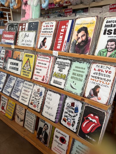 The image displays a retail setting with shelves filled with various decorative metal signs. The signs feature text in Turkish, along with illustrations and photographs of people. Some of the signs have humorous or motivational quotes, while others appear to be related to sports teams or cultural figures. The products are organized on wooden shelves, with some signs still in plastic packaging. In the background, there are glimpses of stuffed animals and other retail items. The overall impression is of a gift shop or souvenir store. The metadata indicates the location as Acıpayam, Turkey.