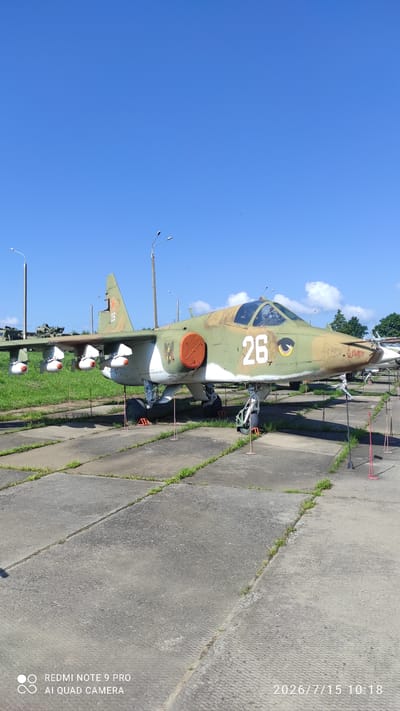 The image shows a parked military aircraft, likely a ground attack jet, outdoors on a concrete pad. The aircraft is predominantly green and brown camouflage with white markings, including the number "26" prominently displayed on its fuselage and tail. Several missiles are attached to pylons under its wings. The aircraft is secured with chocks and orange marker poles are placed around it. In the background, there are streetlights, more military vehicles, and trees. The sky is clear blue with a few white clouds. The concrete pad has grass growing through the cracks. A watermark in the bottom left corner reads "REDMI NOTE 9 PRO AI QUAD CAMERA" and in the bottom right corner, a date and time stamp reads "2026/7/15 10:18". This scene is consistent with an outdoor museum or a preserved military exhibit.