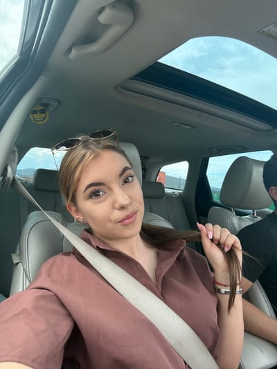 A young woman with long brown hair, wearing a brown collared shirt and sunglasses on her head, is in the passenger seat of a car. She is pulling on a strand of her hair with her right hand, which is adorned with a bracelet and a red string. A seatbelt is across her body. In the background, a man with dark hair is visible in the driver's seat. The car's sunroof is open, revealing a cloudy sky. A yellow smiley face air freshener with sunglasses is hanging from the car's interior. The scene is likely within the city of Aldomirovtsi, Bulgaria.