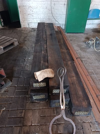 The image depicts the interior of a building, possibly a workshop or storage area, with a cobblestone floor. The scene is dominated by two large, dark, wooden beams positioned side-by-side. Resting on the left beam is a folded piece of light-colored fabric, possibly a rag or cloth. A pair of metal tongs with large circular handles is placed in front of the beams, with its jaws resting on the floor. In the background, a green metal cabinet or door stands to the right, and a white brick wall with peeling paint is visible above it. To the left of the wooden beams, a wooden pallet is partially visible. Several reddish-brown cylindrical objects, possibly pipes or rods, are laid out parallel to the right side of the wooden beams. The overall impression is one of a utilitarian space, potentially related to construction or industrial work. There are no people visible in the image. The location is identified as Vuhlehirsk, Ukraine.
