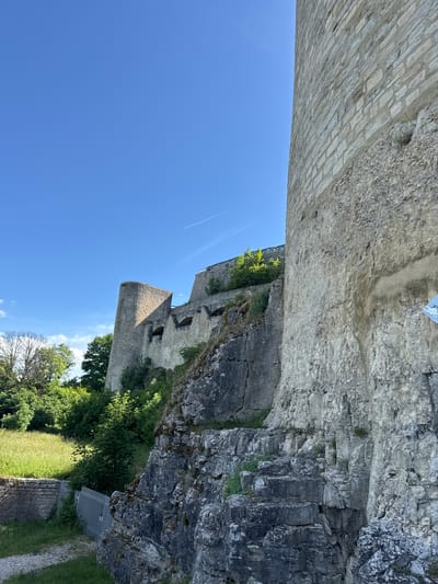 The image shows a view of a stone castle structure under a clear blue sky. The castle walls are made of light-colored stone blocks, with some areas showing rougher, natural rock. Vegetation, including green grass and trees, grows around the base and on some of the ledges of the castle. In the foreground, there is a section of rough, rocky terrain with carved stone steps leading upwards along the castle wall. A metal fence is visible in the lower left corner, bordering a grassy area. A small sign with cartoonish drawings is attached to the castle wall on the right side of the image. The scene appears to be an outdoor historical site, likely part of the Neuffen Castle ruins in Neuffen, Germany, given the context.