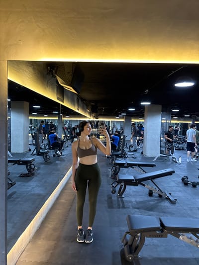 The image shows a woman in a gym, standing in front of a mirror. She is wearing a brown sports bra and olive green leggings, and has a cell phone in her right hand. She is looking at her reflection in the mirror. There are several weightlifting benches in the background and a number of people in the background, including a man in a blue shirt, and another man with a grey t-shirt and black shorts, and a man in a white t-shirt and blue jeans.  The gym is well-lit with fluorescent lights and the floors are tiled. The image is taken in a gym located at latitude 20.56 and longitude -100.432 in El Pueblito, Mexico.