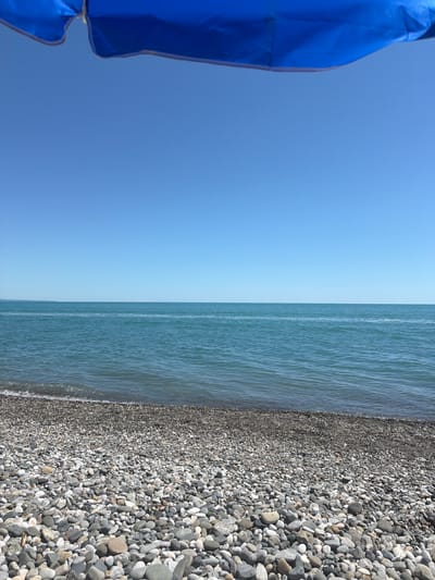 The image depicts a view of the sea from a pebbled beach. In the foreground, a dense collection of smooth, grey and white pebbles covers the beach. A small portion of a blue umbrella is visible in the upper part of the image, casting a shadow over the scene. The sea is a clear, tranquil blue, with gentle waves lapping at the shore. The horizon is visible, with a clear, unblemished blue sky above. The location is identified as Amzara, Abkhazia. There are no people or animals present in the image.