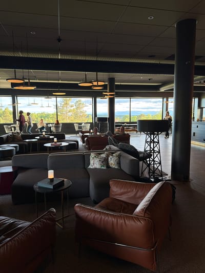 The image depicts the interior of a modern lounge or restaurant with large windows offering a view of a scenic landscape outside, likely in Östersund, Sweden, characterized by rolling hills and trees. Inside, the scene is dimly lit with contemporary pendant lights hanging from the ceiling. The furniture consists of comfortable seating areas with dark grey sectional sofas and rich brown leather armchairs. Small, round side tables with decorative candles and books are placed strategically around the seating. Several people are visible, some standing and conversing near the windows, others seated and relaxing. In the background, a tall, dark pillar and an industrial-style floor lamp are prominent features. The overall ambiance is one of relaxed sophistication and comfort, with the natural scenery providing a striking contrast to the modern interior design.